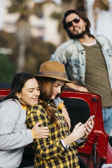 Cheerful women embracing lady with smartphone near car trunk man leaning out from automobile
