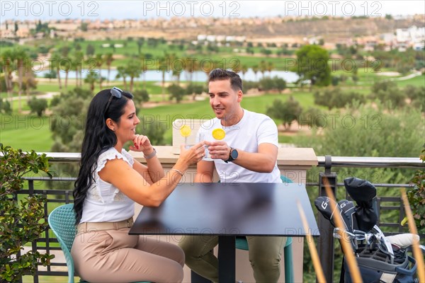 Casual rich couple toasting with cocktails sitting on an outdoor terrace facing a golf court