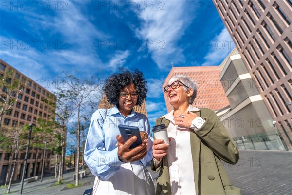 Multi-ethnic businesswomen laughing while using phone outdoors in the street in the financial district