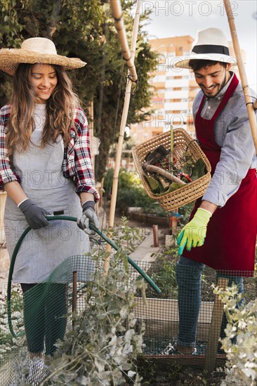 Man guiding smiling female gardener watering plant with green hose