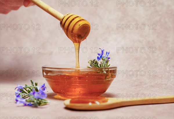 Woman holding a wooden spoon dripping honey over a glass bowl with fresh rosemary branches in bloom