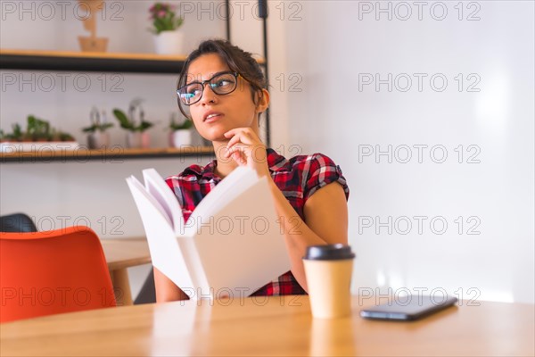 Pensive woman reading a book at home sitting on a table with a coffee cup and mobile phone