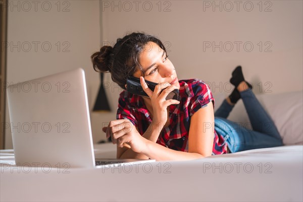 Distracted woman talking to the mobile while working with laptop lying comfortable on an hotel bed
