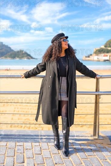 Latin woman in warm clothes posing next tot he beach leaning in a rail in the promenade