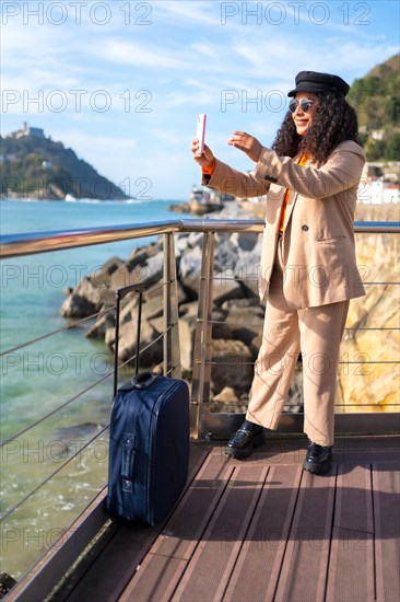 Chic latin woman taking a photo to the sea landscape standing next to luggage in a promenade