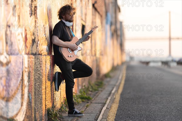 Romantic man hipster wearing sunglasses standing by the wall and playing electric guitar at early sunset. Mid shot