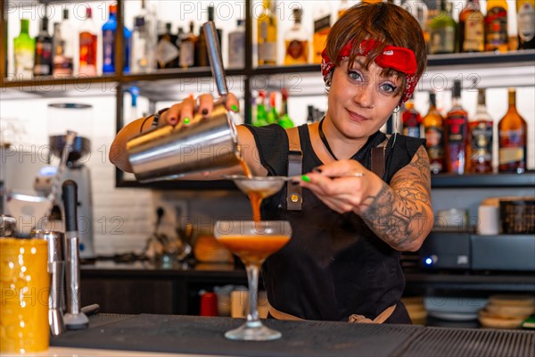 Happy bartender pouring cocktail from the shaker into a cocktail glass in the counter of a bar