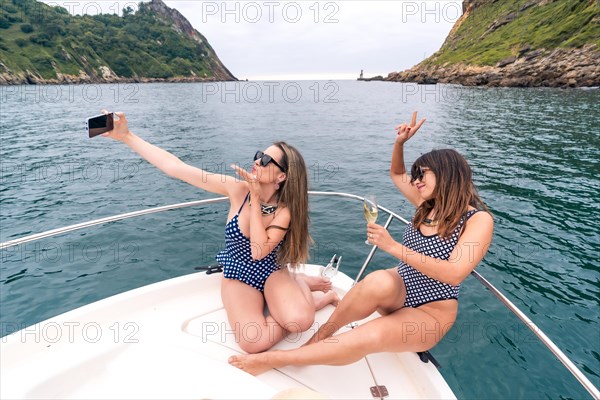 Rich and stylish young women in swimsuit taking selfie while drinking champagne on a ship deck