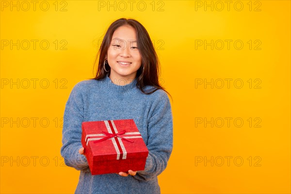 Studio photo with yellow background of a chinese woman giving a present smiling at camera