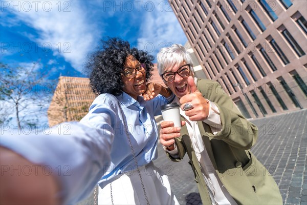 Multi-ethnic businesswomen gesturing with thumb up while taking a selfie in the street in the morning