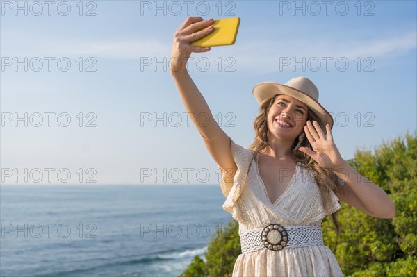 Horizontal photo of a blonde beauty woman in summer clothes waving while taking a selfie next to the sea