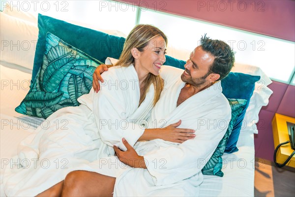 Newlywed couple sitting on the bed of a luxury hotel embracing and wearing white bathrobe