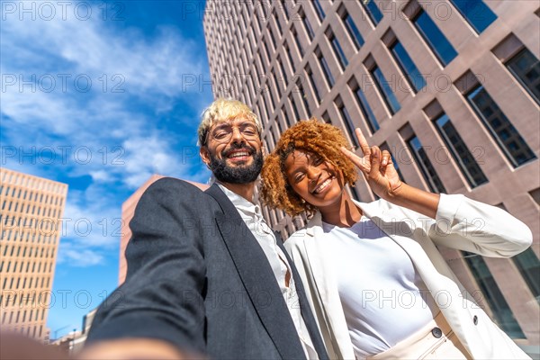 Low angle view portrait of two successful business colleagues smiling and taking a selfie outdoors in a sunny day in the city