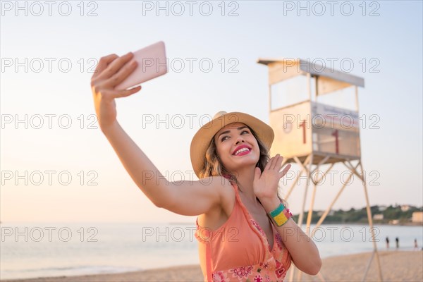 Woman with summer clothes waving at camera taking a selfie with the mobile on the beach during sunset