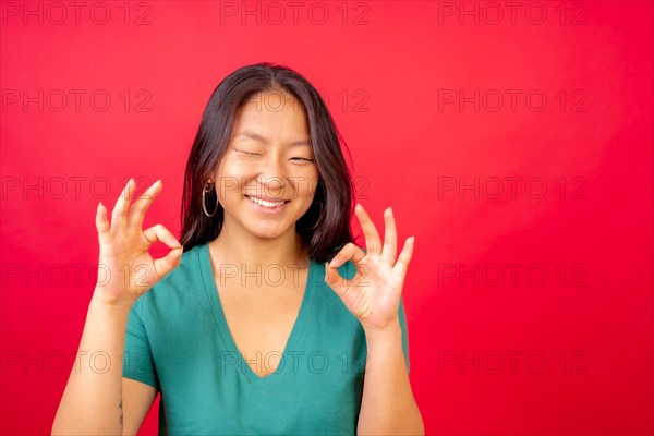 Studio photo with red background of a young chinese woman gesturing ok with hands and winking