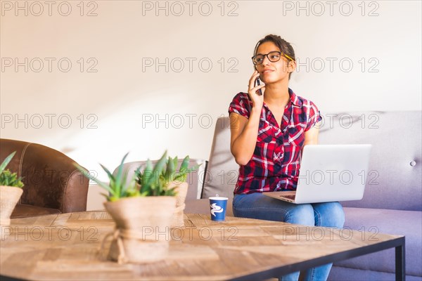 Woman having a conversation with mobile while working at home using laptop sitting on a sofa