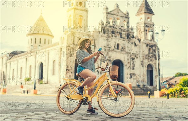 Happy girl in hat riding a bicycle and texting with the phone. Happy tourist girl in hat on bicycle using phone on the street. Granada