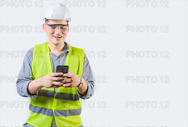 Handsome engineer holding phone isolated. Cheerful builder engineer texting phone on isolated background. Young engineer in vest and helmet using cell phone