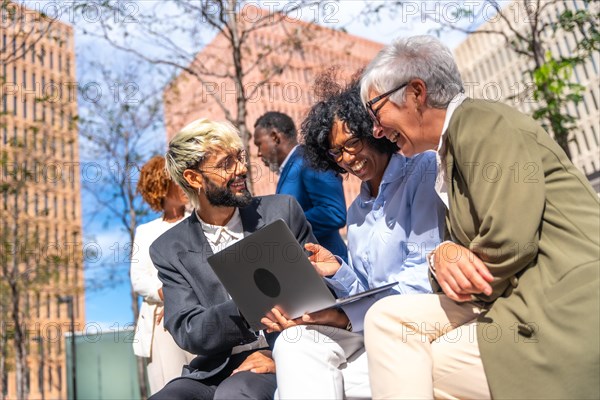 Happy business team using laptop sitting outdoors in a bench next to a financial building