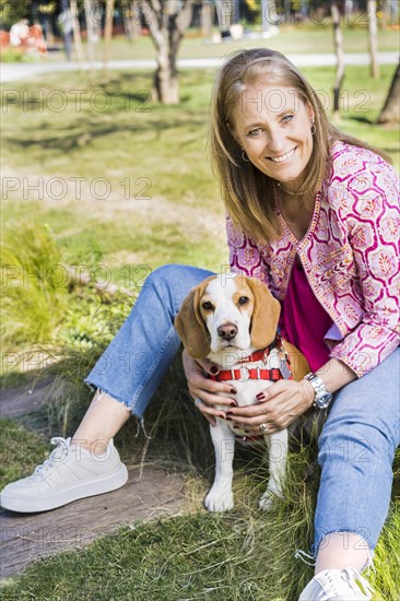Happy blond mid adult woman smiling for camera and hugging a Beagle dog while sitting on green grass at the park