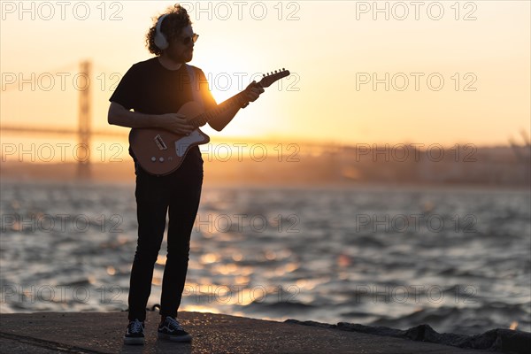 A man stands at the waterfront and playing guitar at bright orange sunset. Mid shot