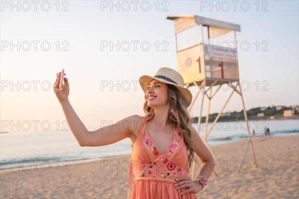 Happy woman with summer dress and hat taking a selfie on the beach at sunset