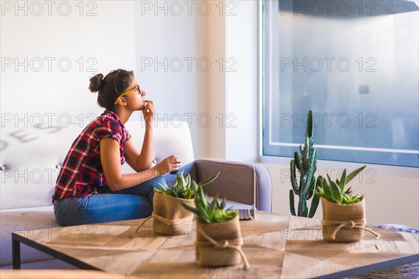 Dreamy woman looking through the window at home while using laptop and drinking coffee