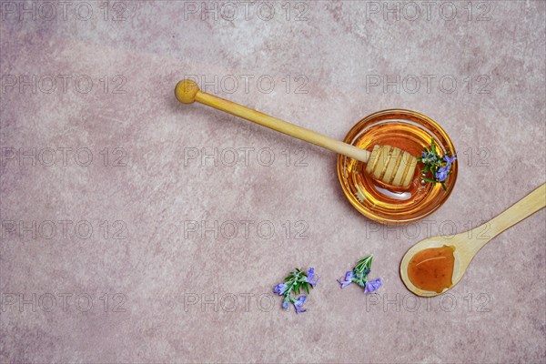Top view of natural rosemary honey in a glass bowl with a wooden spoon and fresh rosemary branches in blossom