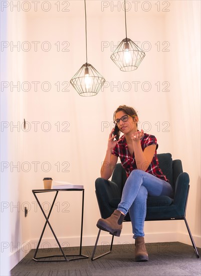 Vertical photo of a woman talking to the mobile sitting with a coffee at home in a cozy room