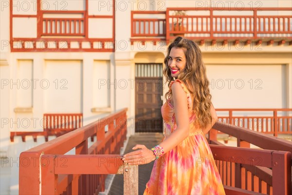 Pretty woman with long dress turning to smile at camera during sunset in a coastal bridge