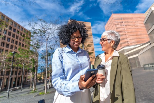 Multi-ethnic businesswomen smiling while using phone outdoors in the street in the financial district