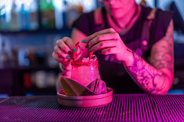 Close-up photo of a female alternative bartender decorating a margarita cocktail with devil horns in the counter of a night bar with neon lights