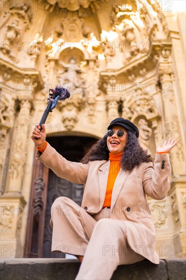 Vertical low angle photo of a female latin influencer posing while recording a video using phone and microphone sitting in a monument in winter