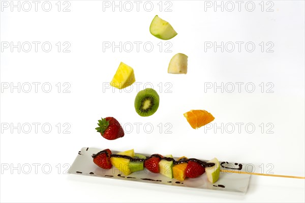 Skewer of assorted fruits covered with chocolate with pieces of fruit falling isolated on a white background