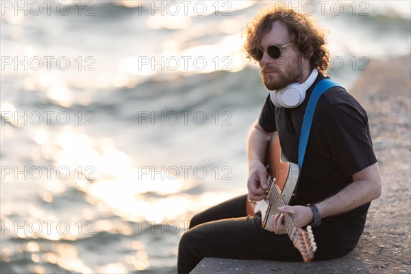 Man hipster playing guitar at the waterfront. Mid shot