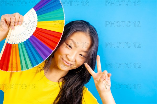 Studio photo with blue background of a chinese woman using a rainbow colored folding fan gesturing peace