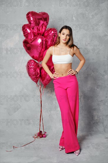 Cheerful young woman in wide bright pink trousers and white tube top posing with arms on waist next to balloons in studio