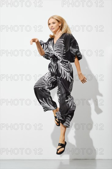 Playful blonde woman in patterned overall jumping indoors