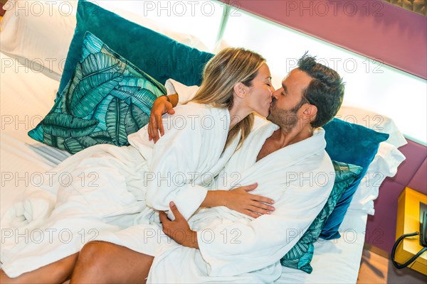 Couple during a honeymoon in a luxury hotel room kissing sitting on a bed wearing white bathrobe