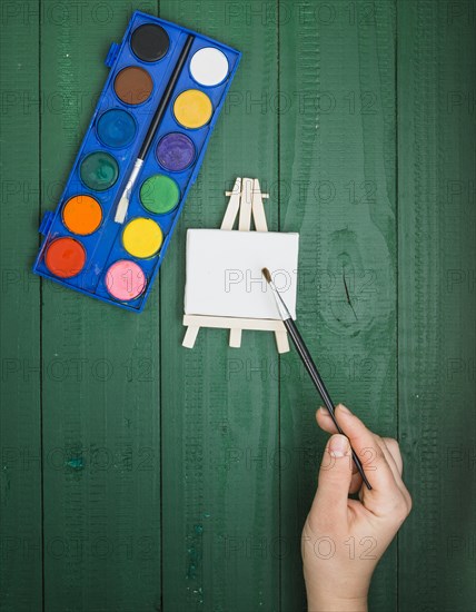 Elevated view hand holding paint brush mini easel watercolor palette