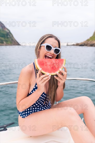 Vertical photo of a young woman n swimsuit eating watermelon on the ship deck