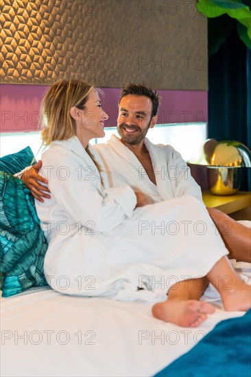 Vertical photo of a happy couple sitting on a bed of a luxury hotel dressing bathrobe