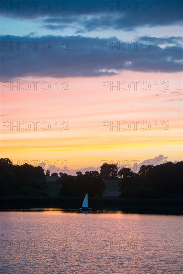 A sailing boat with a white sail lies on the water at sunset against the glowing orange and blue evening sky with dark clouds