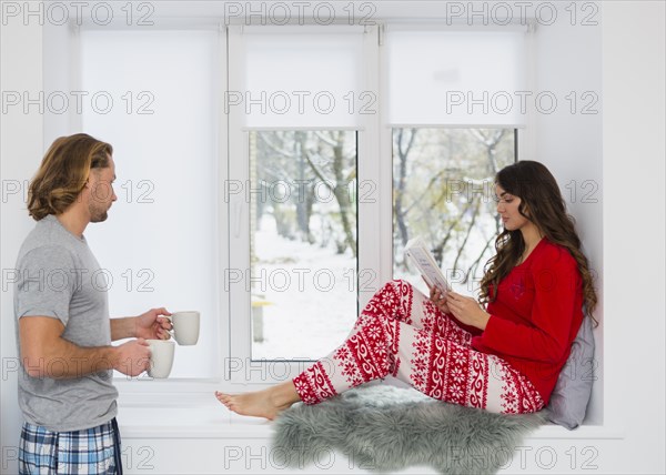 Man bringing coffee mug woman sitting window sill reading book