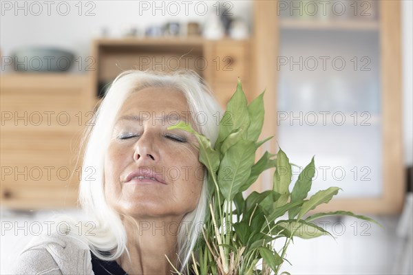 Elderly woman with long hair and closed eyes snuggles up to houseplant at home