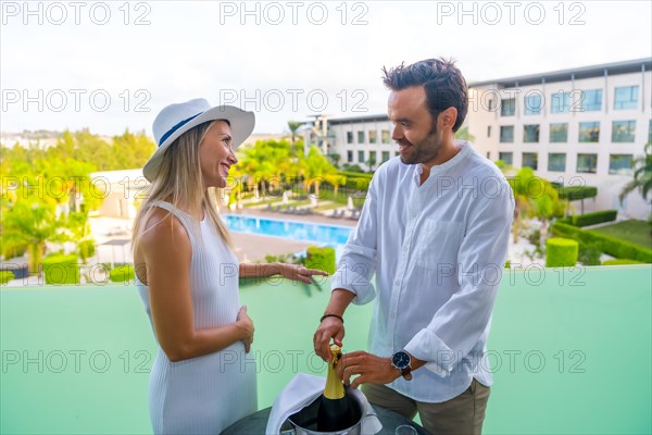 Couple opening a bottle of champagne in an hotel terrace with beauty views of a pool