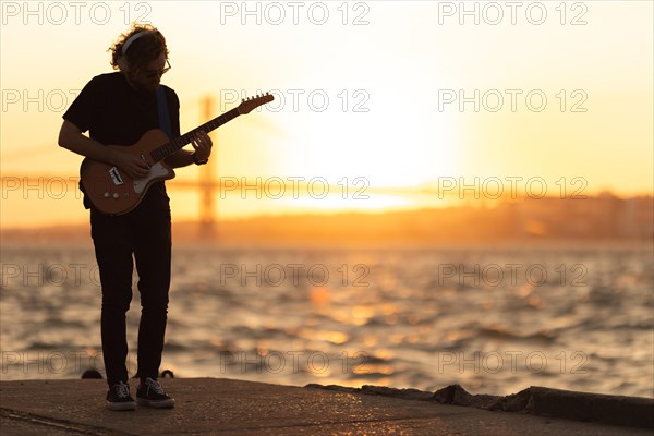 A man stands at the waterfront and playing guitar at bright sunset. Mid shot