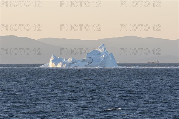 An iceberg floats in the rising morning sun against the silhouette of the mountains of the Greenland coast. Disko Bay