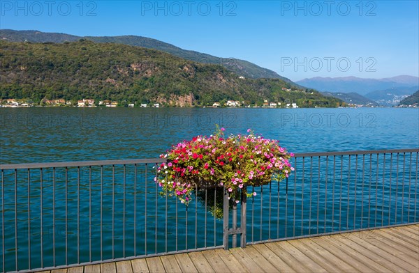 Elevated Walkway with Flowers on the Railing on Lake Lugano with Mountain in a Sunny Summer Day in Porto Ceresio