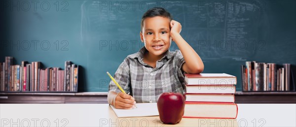 Happy young hispanic school boy at desk with books and apple in front of chalkboard